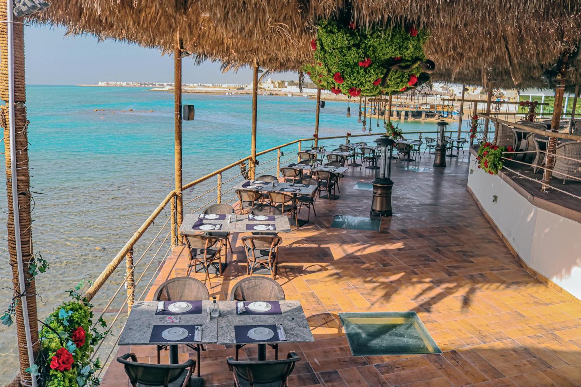 Lagoona Beach Luxury Resort And Spa Manáma Kültér fotó The photo shows a seaside restaurant with a spacious outdoor dining area. It features several tables set with plates and utensils, surrounded by lush greenery and decorative plants. The flooring is made of brown tiles, and there are thatched roofs pr