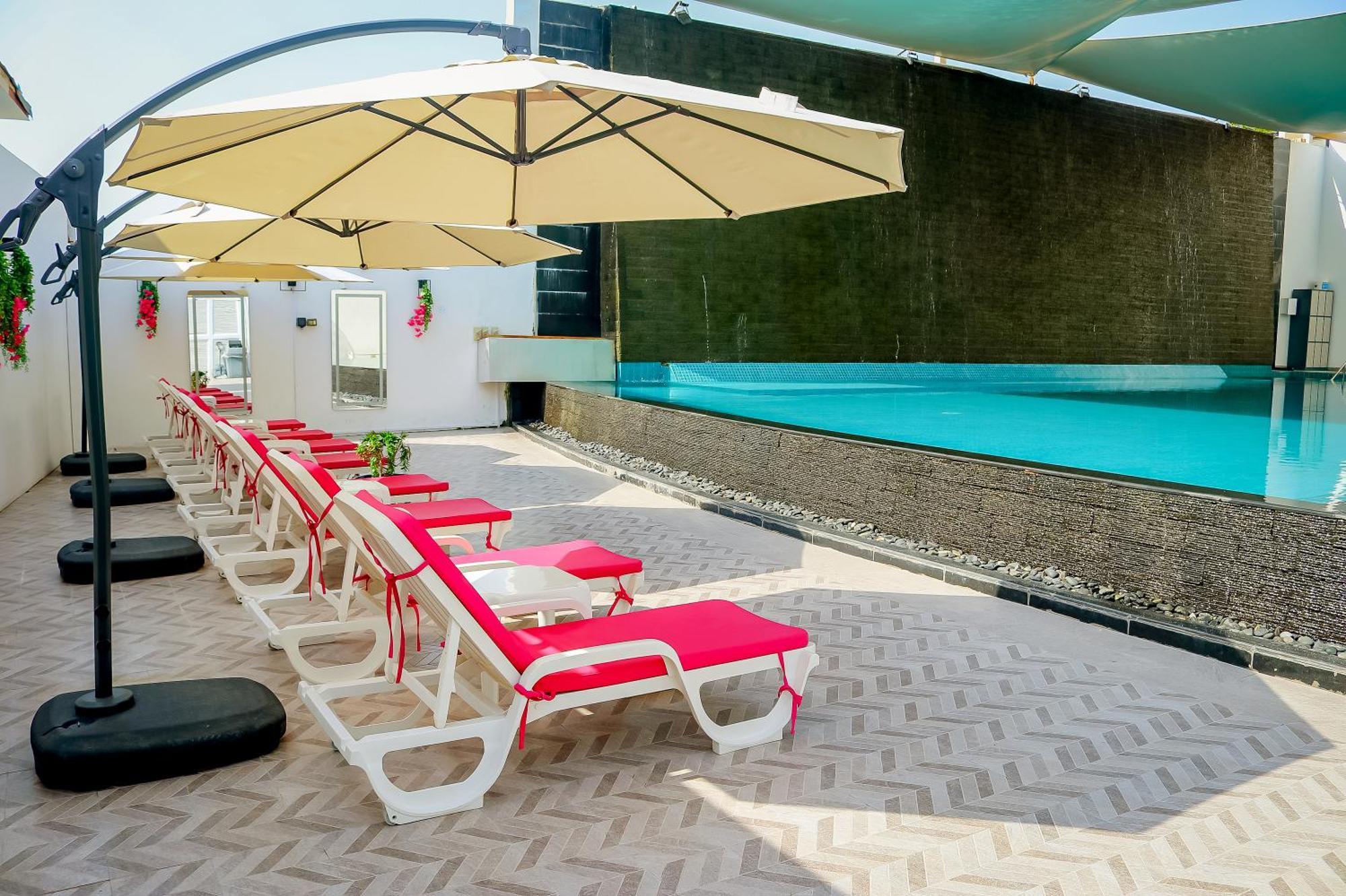 Lagoona Beach Luxury Resort And Spa Manáma Kültér fotó The photo shows a sunny poolside area with several lounge chairs arranged in a row. Each chair features red cushions and is positioned under large, beige umbrellas for shade. In the background, there is a beautiful swimming pool with clear blue water
