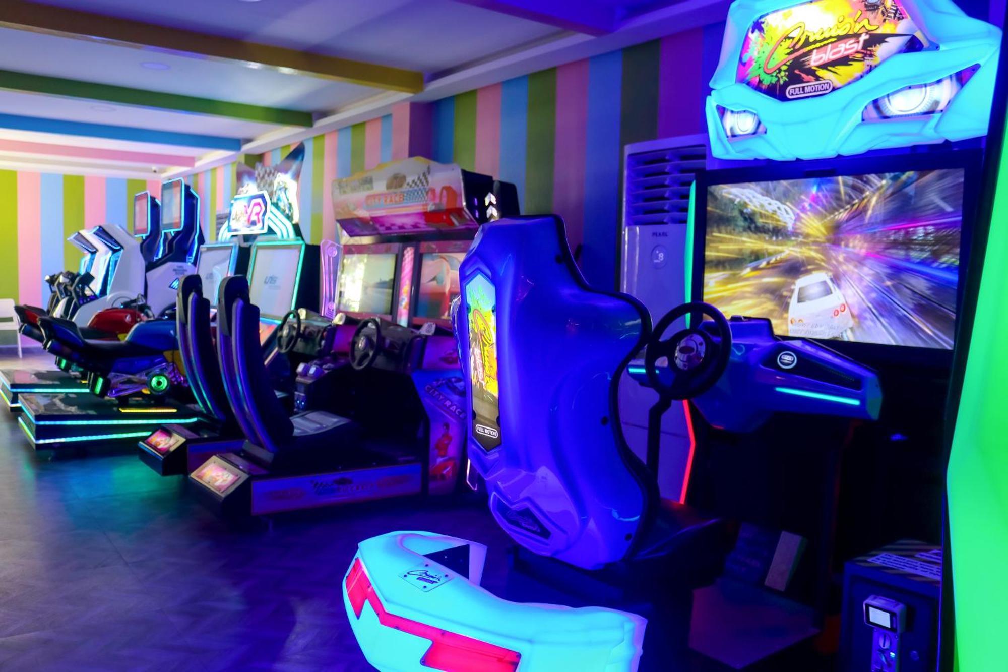 Lagoona Beach Luxury Resort And Spa Manáma Kültér fotó The photo shows an arcade area filled with various arcade machines. The setting is illuminated with colorful neon lights, creating a vibrant atmosphere. There are several racing game machines with steering wheels and seats, each accompanied by screen