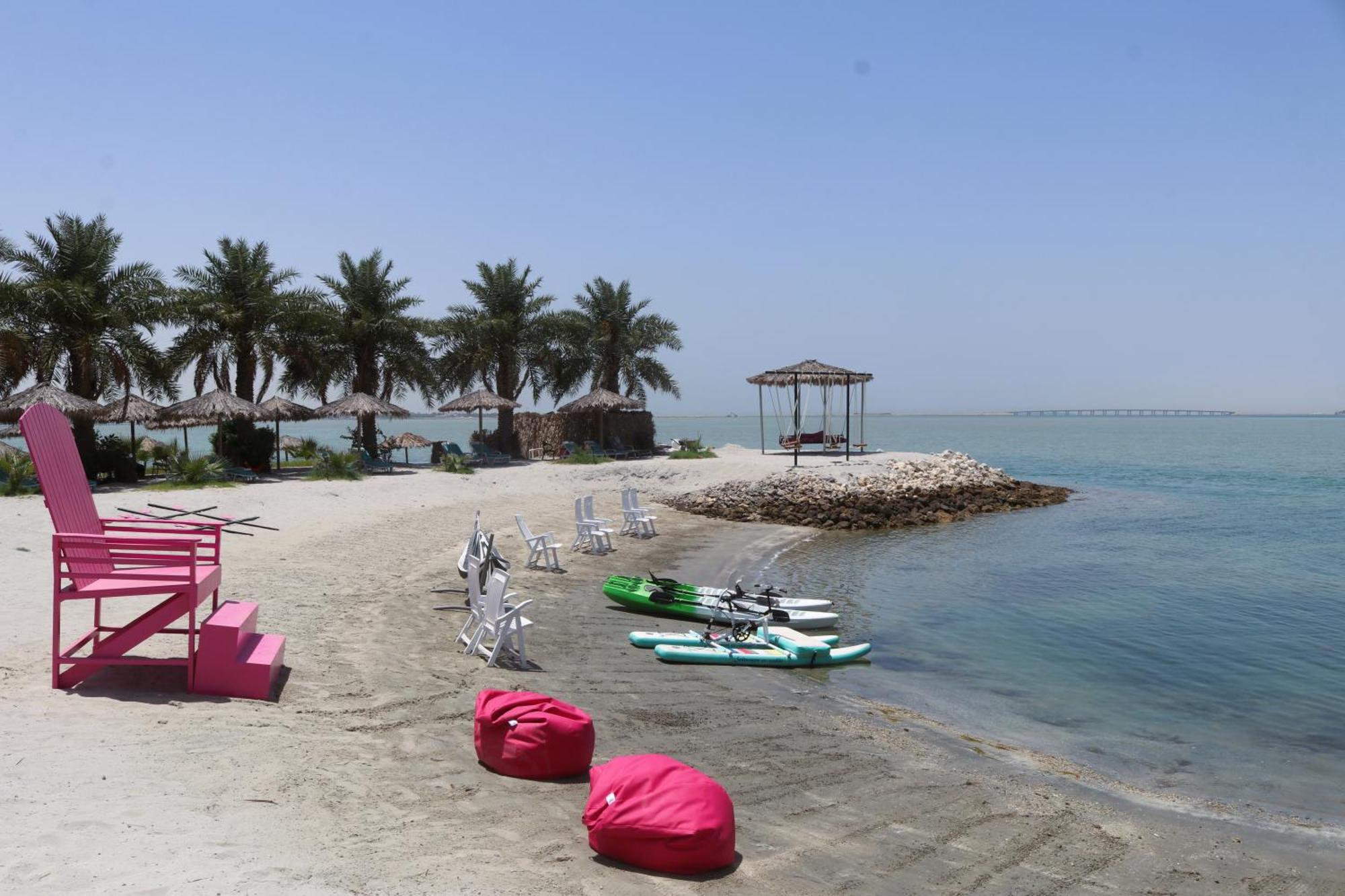 Lagoona Beach Luxury Resort And Spa Manáma Kültér fotó The photo depicts a serene beach scene with clear blue water and a sunny sky. In the foreground, there are colorful pink bean bags and a large pink chair on the sand. A few white chairs and loungers are positioned near the water's edge. To the side, 