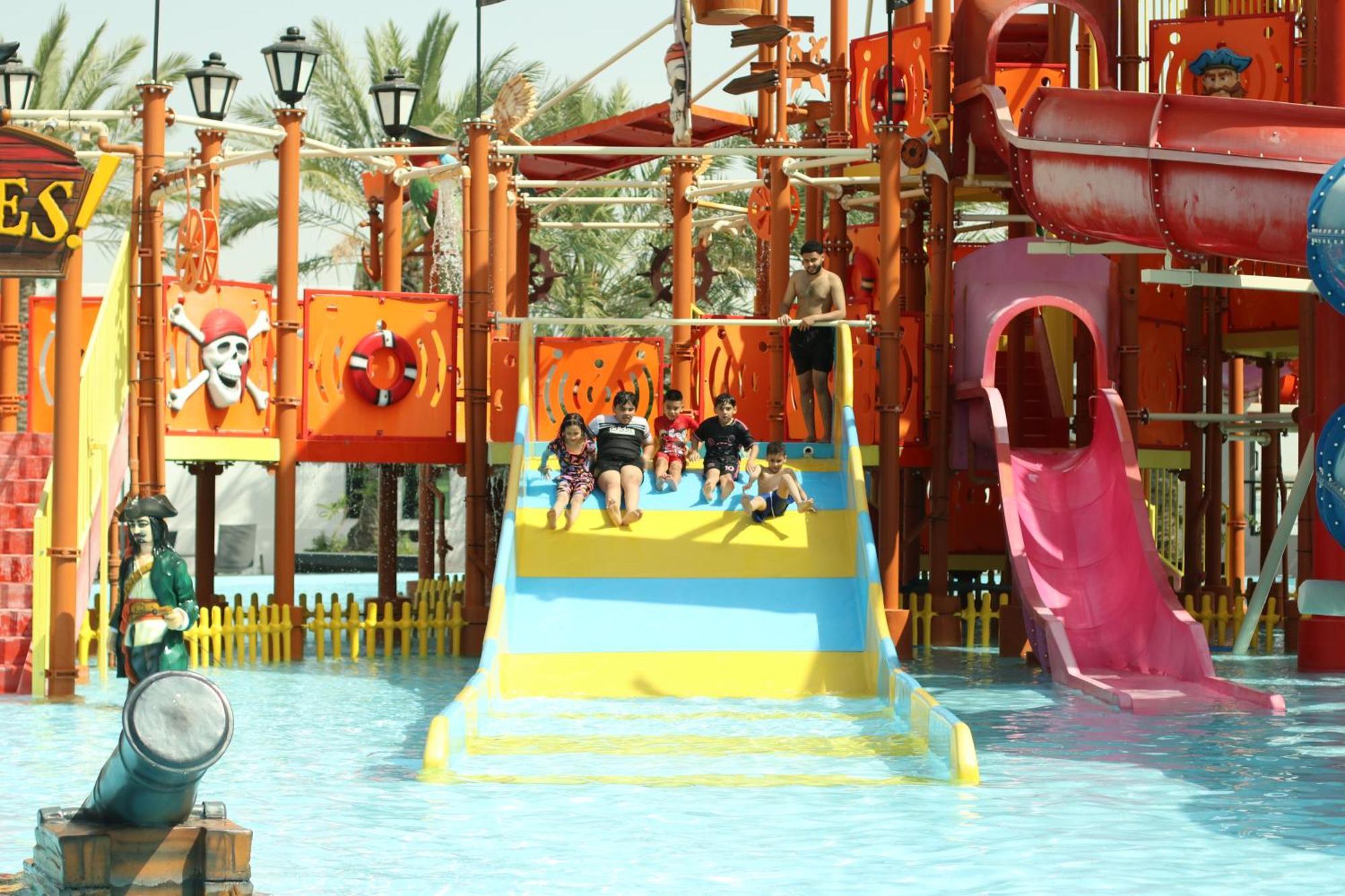 Lagoona Beach Luxury Resort And Spa Manáma Kültér fotó The photo shows a colorful water park scene featuring a play area with multiple slides. There are children enjoying themselves, sliding down bright yellow and pink slides into a pool of water. The structure has pirate-themed decorations, including a 