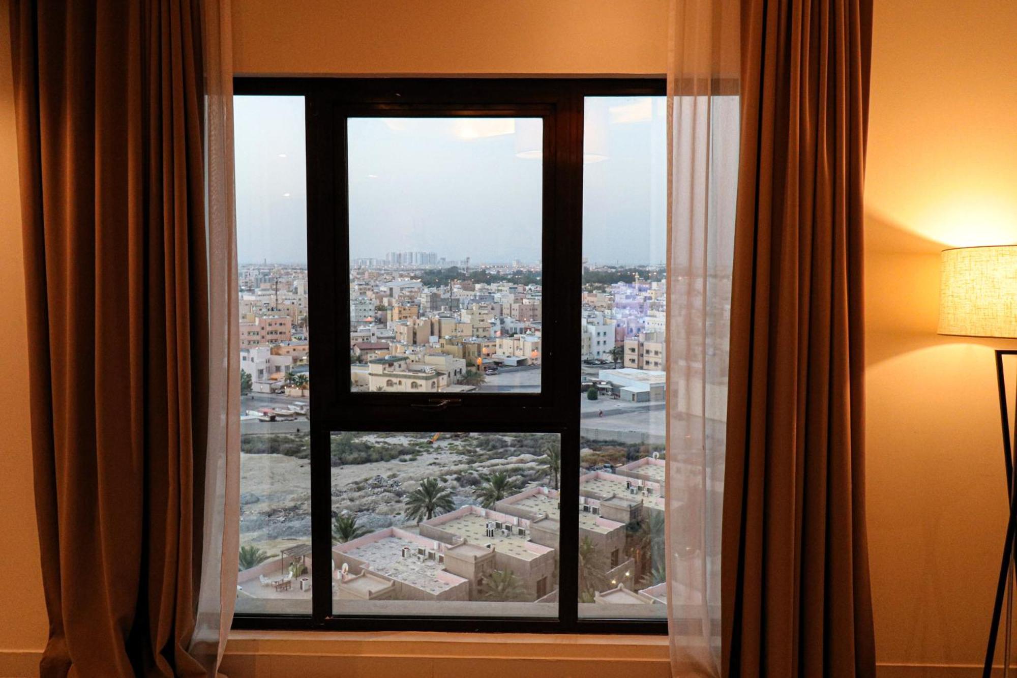 Lagoona Beach Luxury Resort And Spa Manáma Kültér fotó The photo shows a window with a black frame and sheer curtains. Through the window, there is a view of a cityscape, displaying a densely populated area with various buildings and rooftops. The sky appears slightly overcast, suggesting a muted lightin