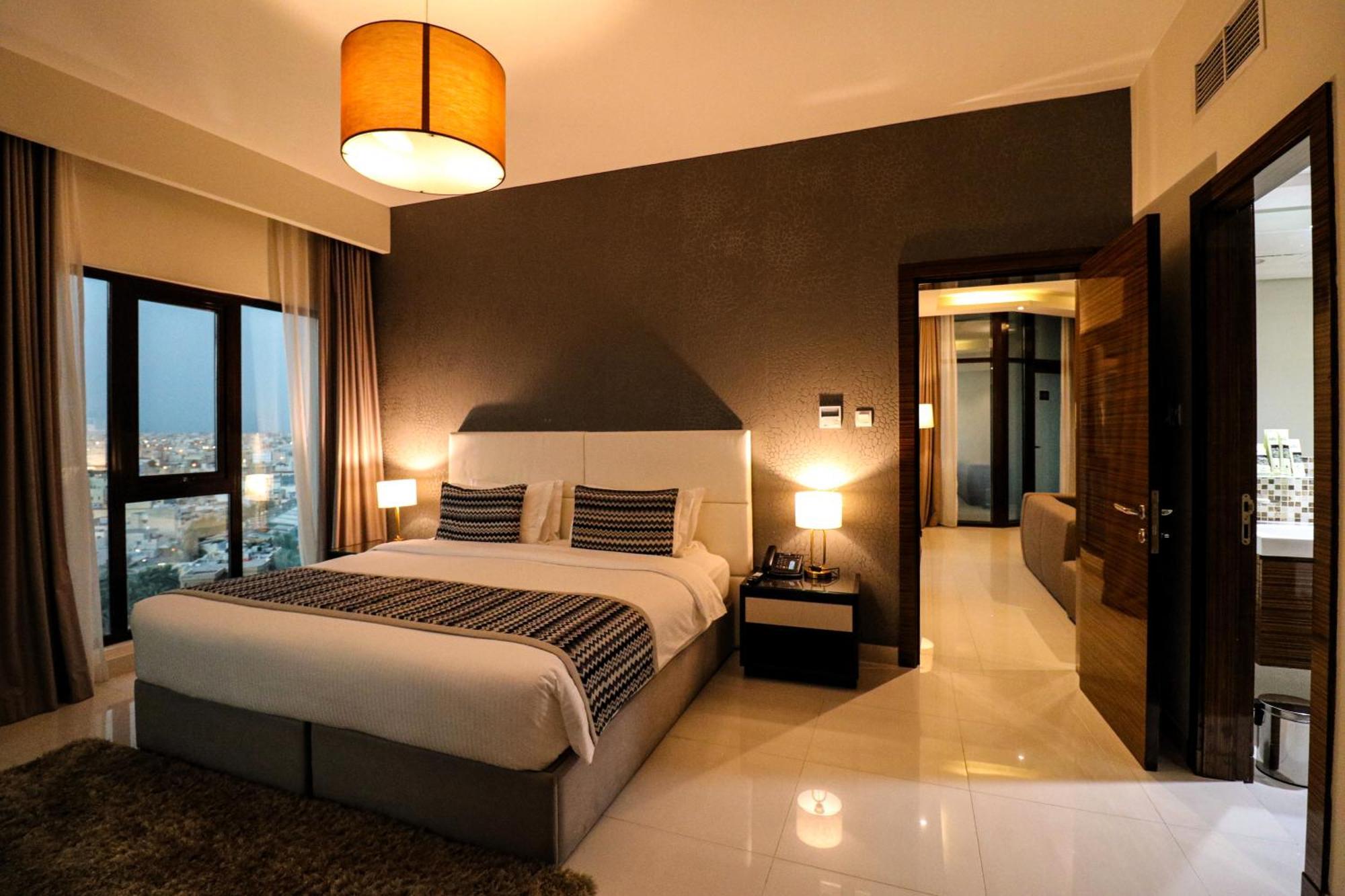Lagoona Beach Luxury Resort And Spa Manáma Kültér fotó The photo depicts a modern hotel room or suite. It features a large bed with a white and gray color scheme, accented with decorative pillows. On either side of the bed, there are sleek bedside lamps providing warm lighting. The walls are a dark color