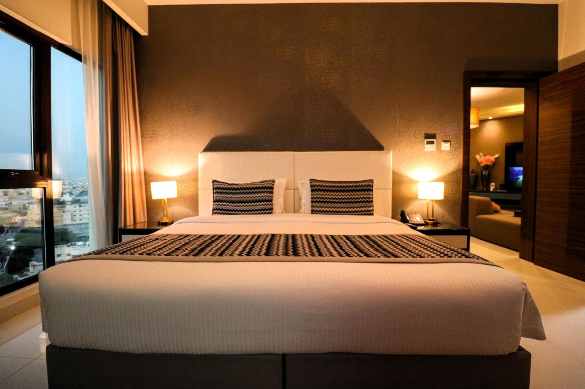 Lagoona Beach Luxury Resort And Spa Manáma Kültér fotó The photo depicts a modern hotel room featuring a large bed with a white comforter and two decorative pillows. The headboard is light-colored, and the walls are painted a dark hue. On either side of the bed, there are bedside tables with lamps. The r