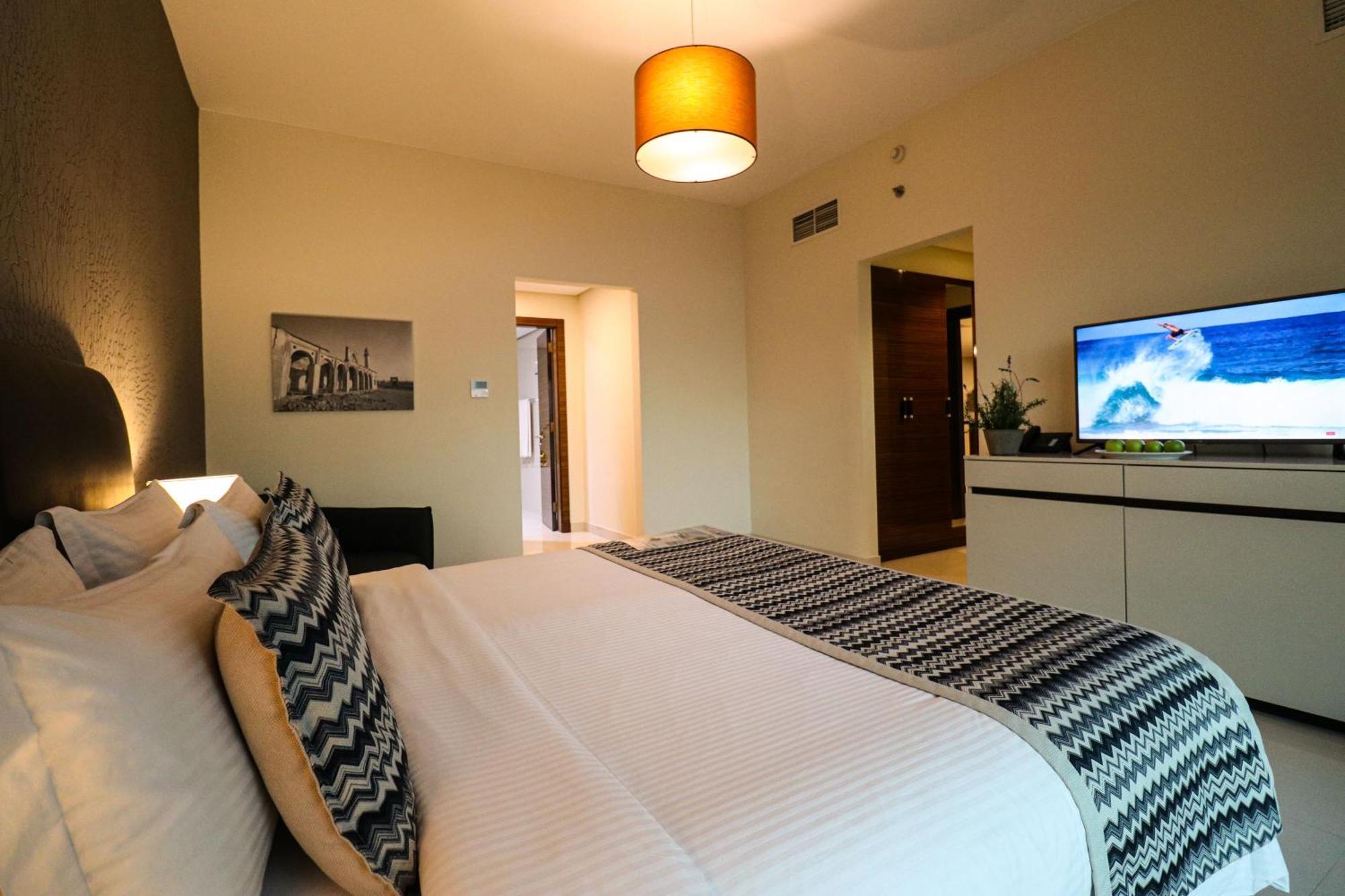 Lagoona Beach Luxury Resort And Spa Manáma Kültér fotó The photo shows a well-decorated hotel room. There is a large bed with white linens and decorative pillows featuring a zigzag pattern. On the wall beside the bed is a piece of artwork, and there is a modern television on a white furniture unit opposi