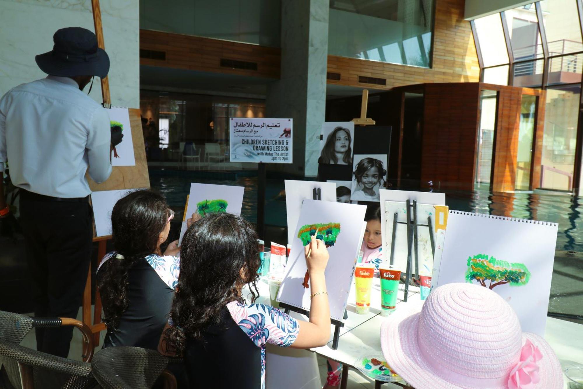 Lagoona Beach Luxury Resort And Spa Manáma Kültér fotó The photo shows a group of children and an adult engaged in an art activity near a pool area. The children are seated at a table, painting on canvases or paper, with a focus on trees, as indicated by the green paint they are using. An adult, wearing 