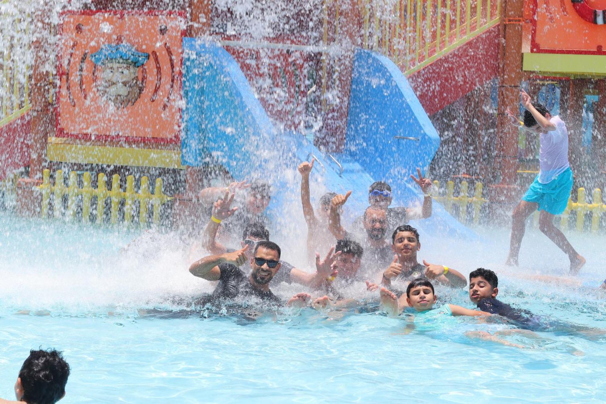 Lagoona Beach Luxury Resort And Spa Manáma Kültér fotó The photo shows a lively scene at a water park or pool area. Several people, including adults and children, are enjoying themselves in the water, splashing and posing for the camera. Water is cascading down, creating a refreshing atmosphere. In the b