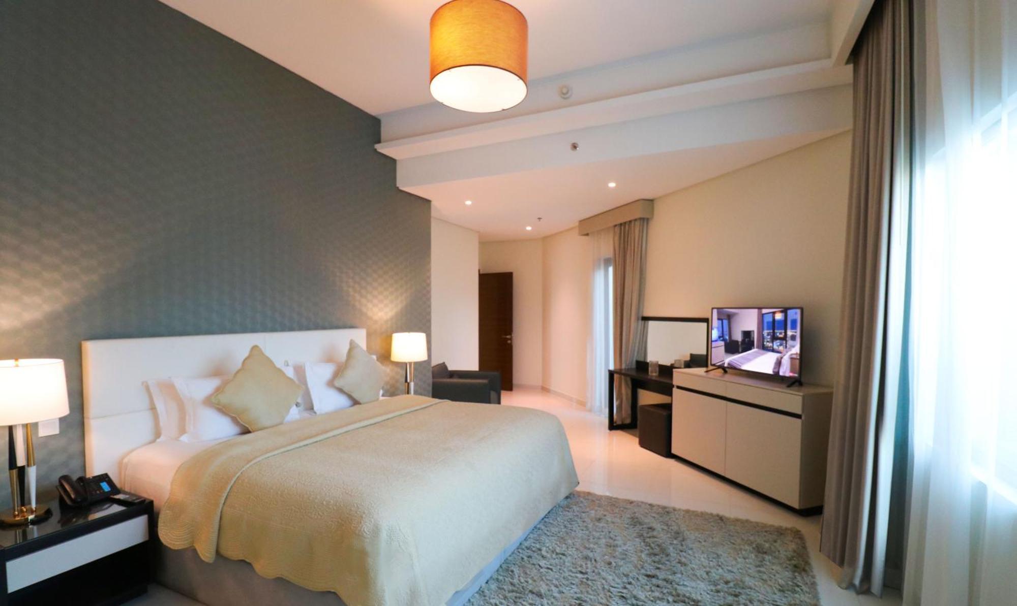 Lagoona Beach Luxury Resort And Spa Manáma Kültér fotó The photo shows a modern hotel room featuring a large bed with a light-colored blanket and decorative pillows. The walls are painted in a subtle shade, and there is a textured accent wall behind the bed. On one side, a bedside lamp provides warm ligh