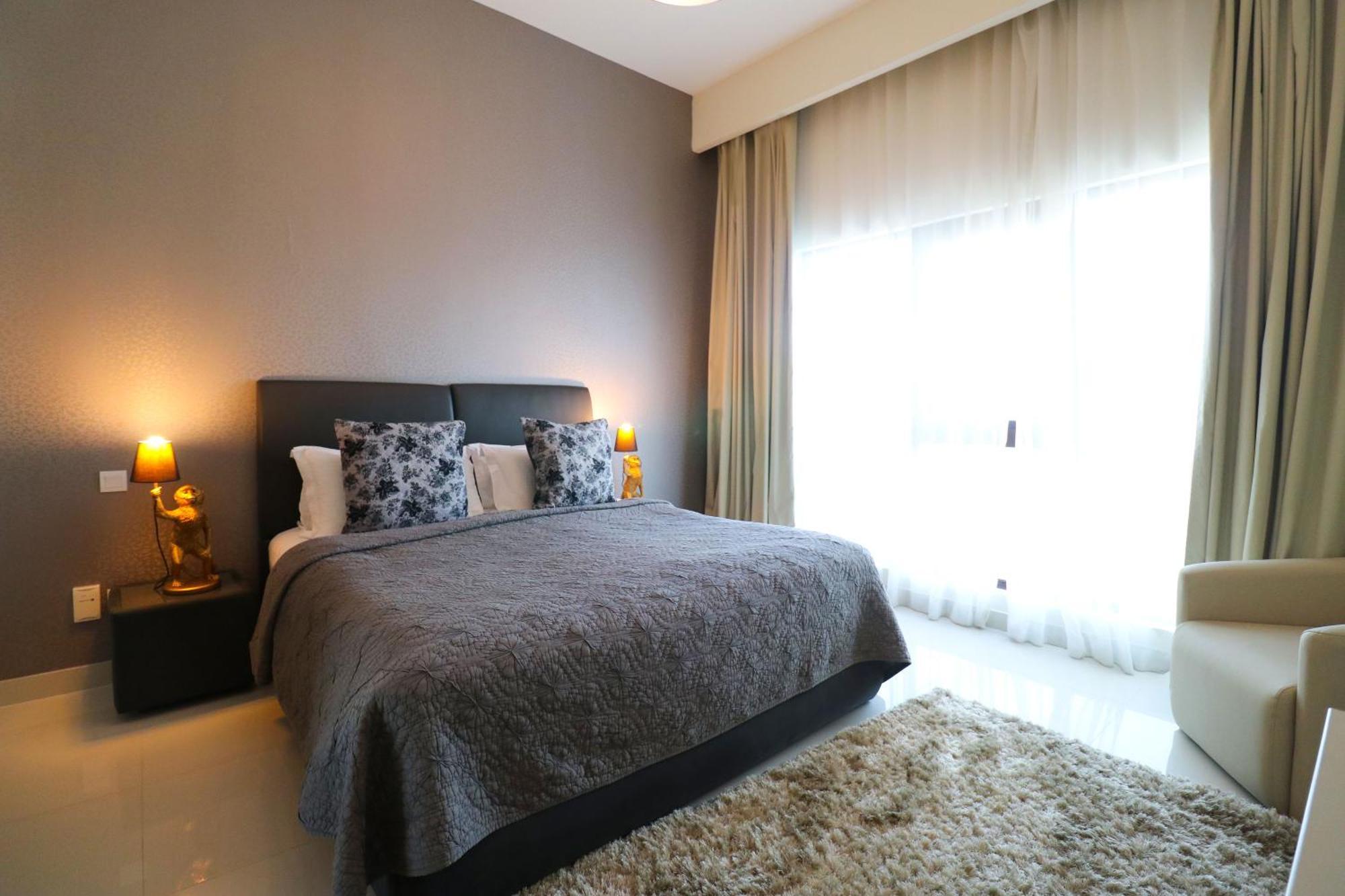 Lagoona Beach Luxury Resort And Spa Manáma Kültér fotó The photo shows a stylish bedroom with a modern design. It features a large bed with a dark gray bedspread and decorative pillows. On either side of the bed are two wooden nightstands, each with a lamp. There is a large window covered with light, flo